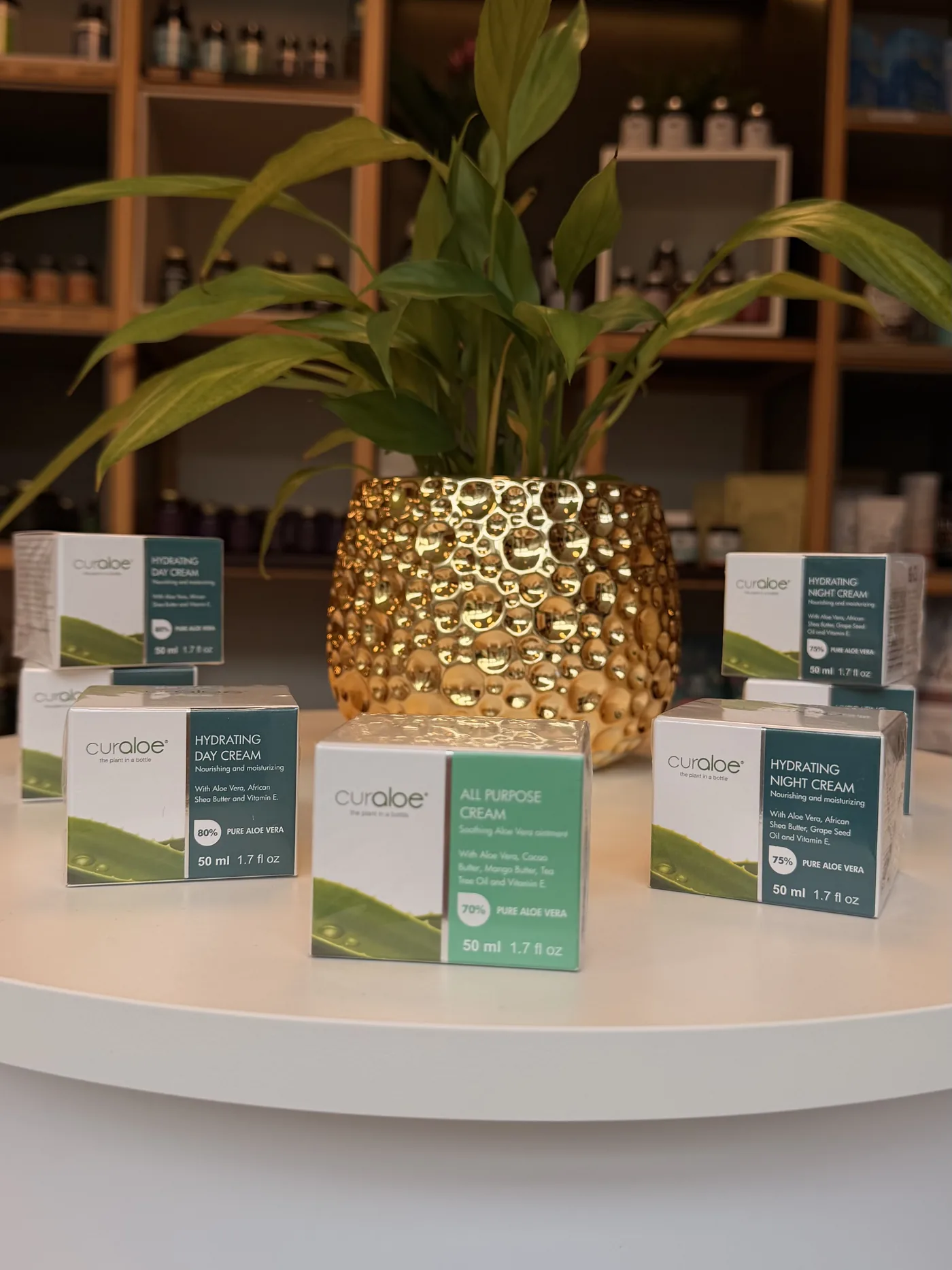 Přírodní kosmetika s aloe vera na kulatém stolku se zlatým květináčem, v pozadí regály prodejny